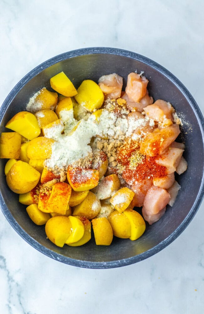 A bowl containing chopped yellow potatoes, diced raw chicken, and various seasonings on a white marble surface.