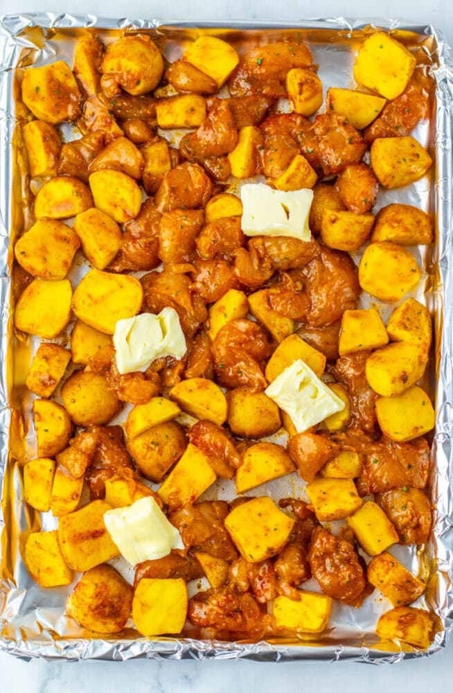 A foil-lined baking sheet with diced potatoes and seasoned chicken pieces, topped with four pats of butter, ready to be cooked.