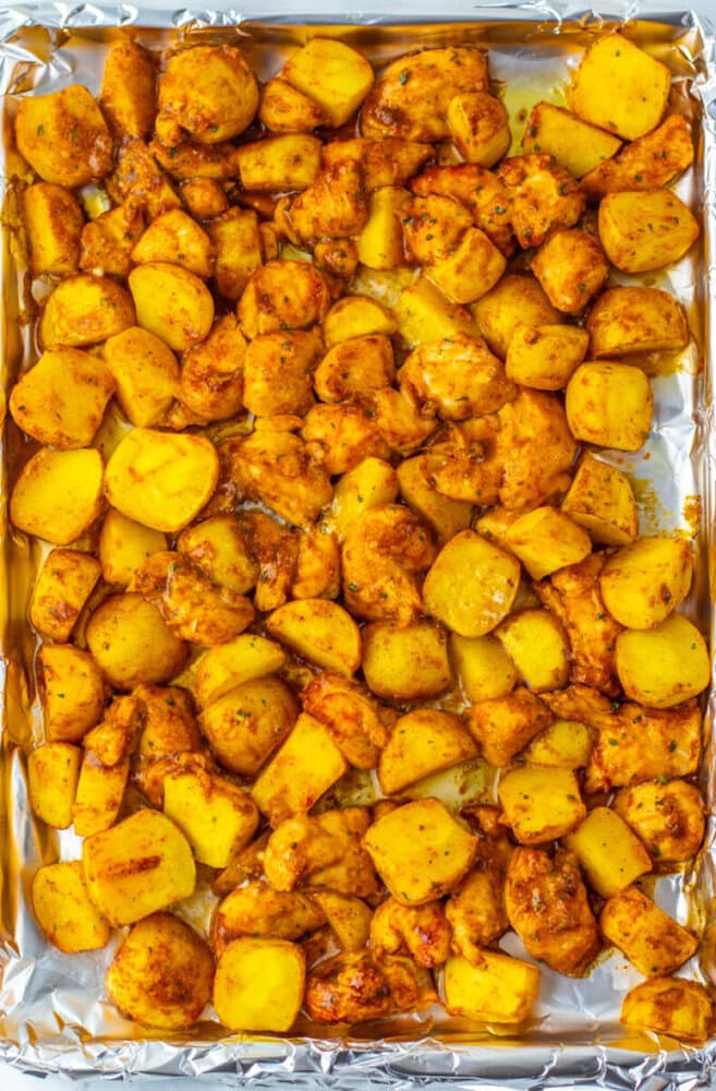 A foil-lined baking sheet filled with evenly spread, roasted yellow potatoes and chicken pieces, seasoned with spices.