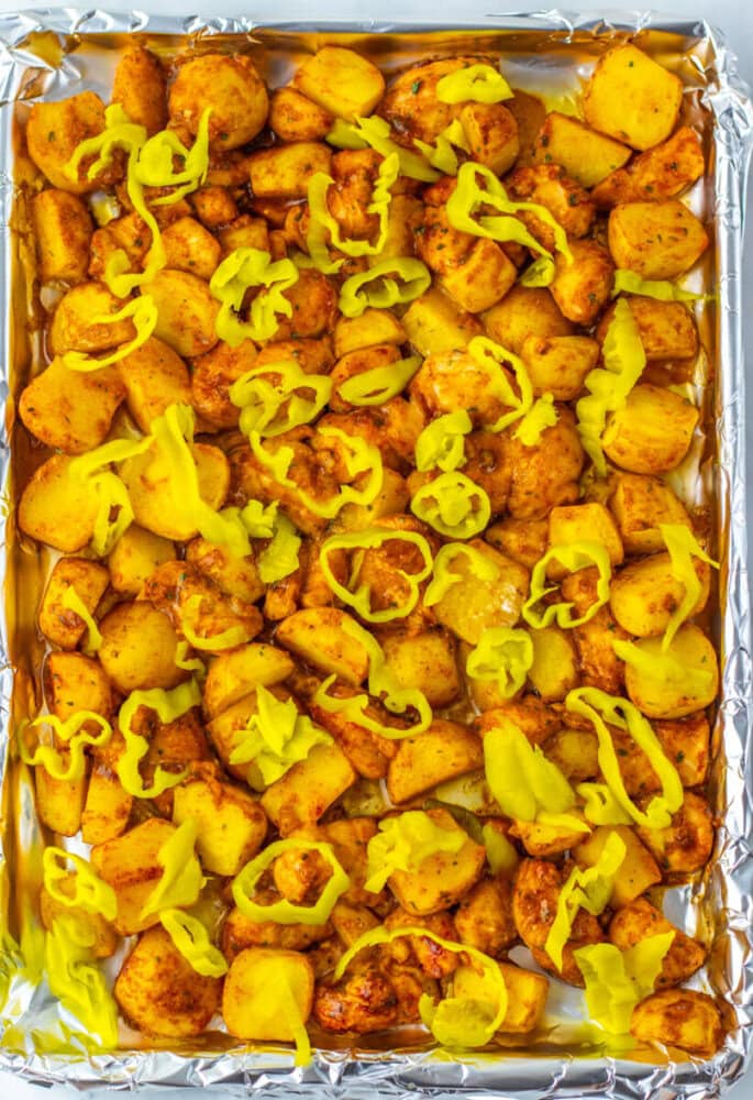 A foil-lined baking sheet filled with roasted potatoes and chicken pieces, topped with sliced yellow peppers.