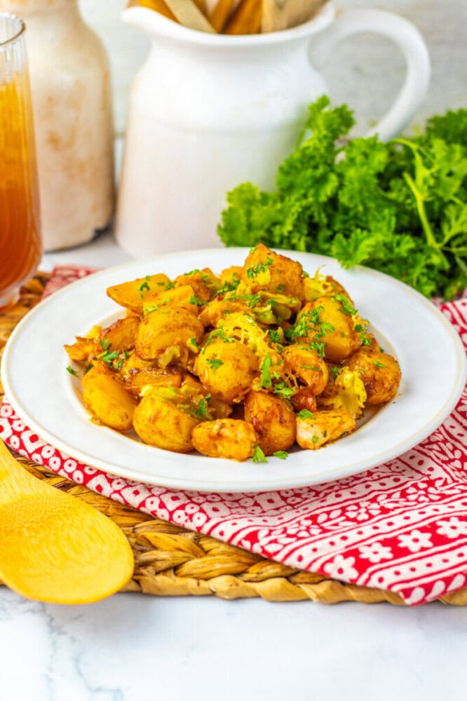 A white plate of seasoned, roasted potatoes garnished with chopped parsley, placed on a red-patterned napkin with fresh parsley and a drink in the background.