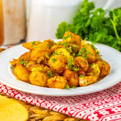 A white plate of seasoned, roasted potatoes garnished with chopped herbs, set on a red patterned cloth near a wooden spoon, with parsley and a drink in the background.