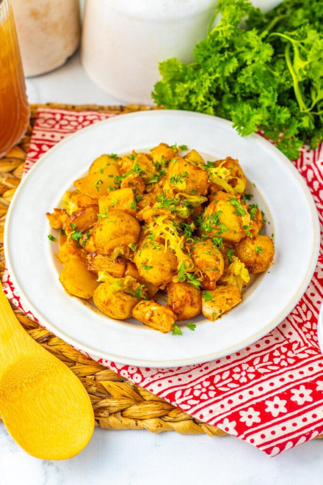 A plate of cooked potatoes and chicken garnished with chopped parsley, placed on a white plate with a red patterned napkin underneath. Fresh herbs and a wooden spoon are nearby.