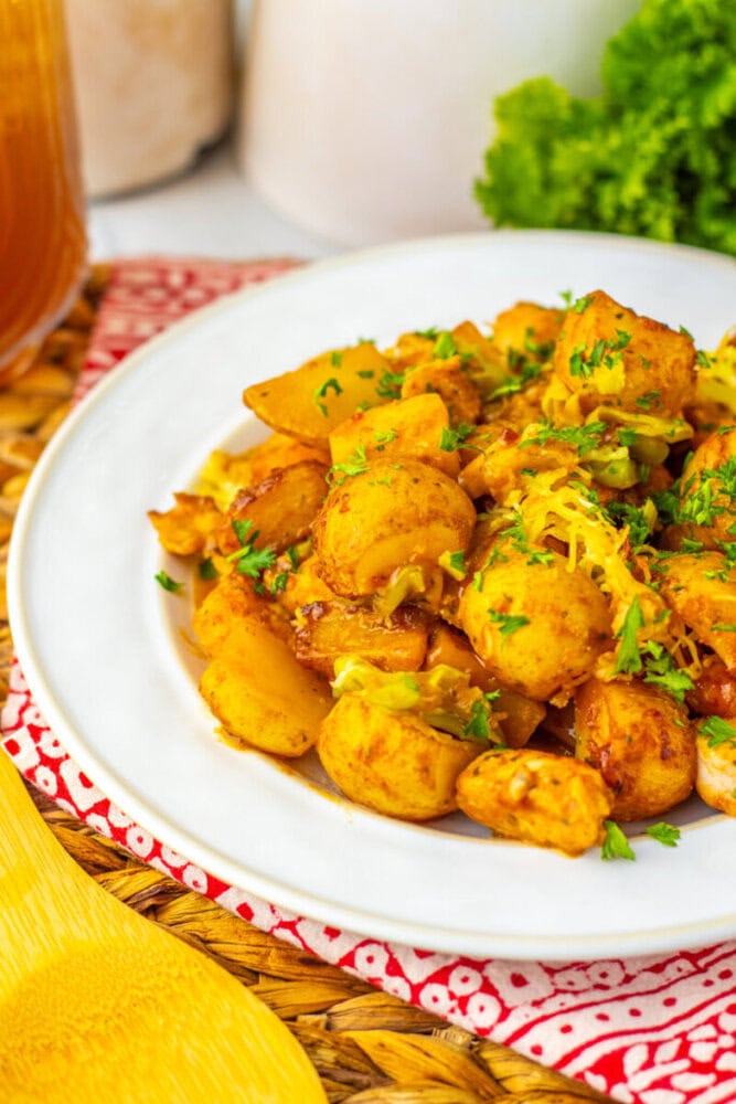 A white plate filled with cooked, seasoned potatoes garnished with chopped herbs and shredded cheese, set on a red-patterned cloth.