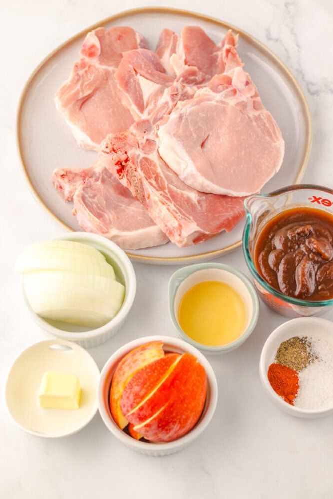 Raw pork chops on a plate with sliced onion, apple slices, butter, apple cider vinegar, spices, and a measuring cup of barbecue sauce arranged on a white surface.