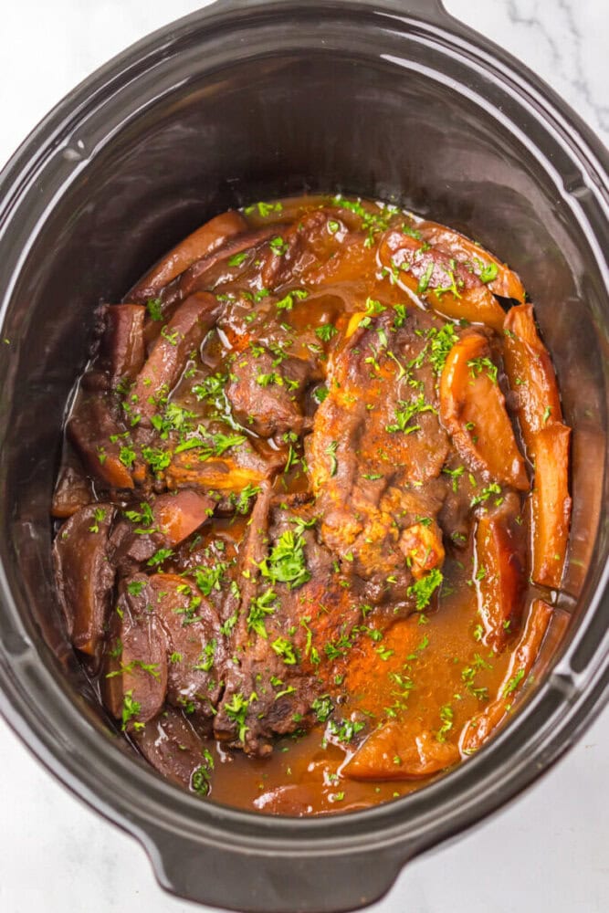 Beef pot roast with sliced carrots and sauce, garnished with chopped parsley, in a black slow cooker on a white surface.
