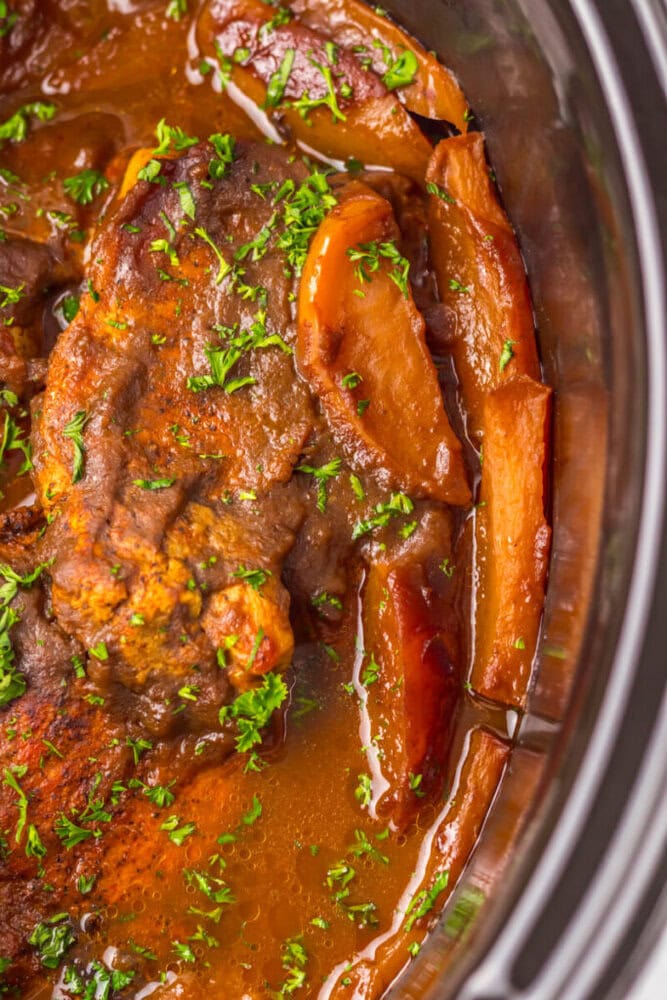 Close-up of a cooked beef roast with sliced apples and chopped parsley in a rich brown sauce inside a slow cooker.
