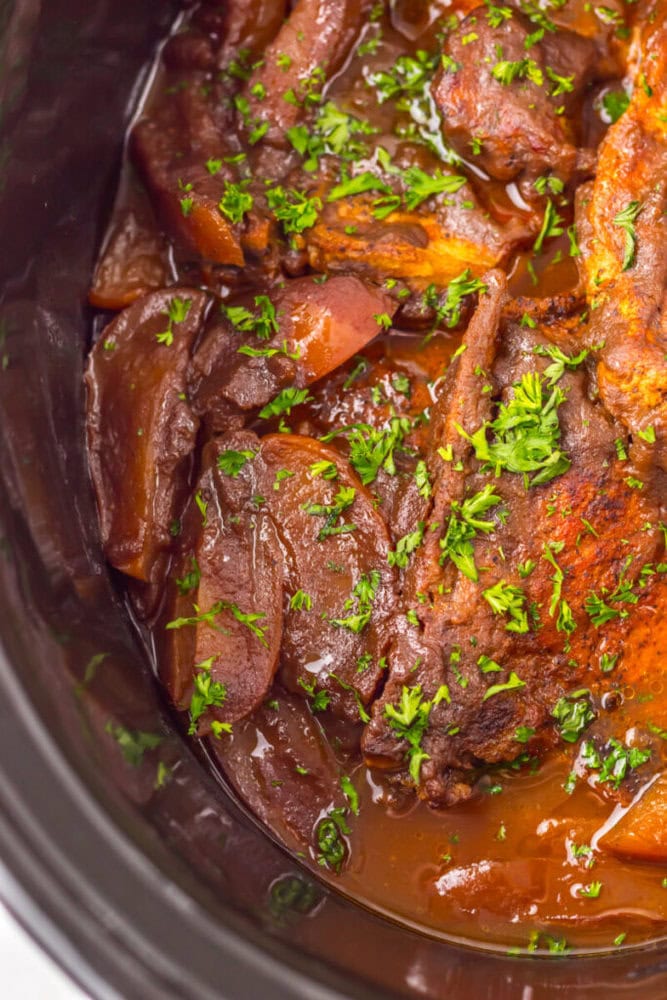 Close-up of slow-cooked meat with carrots and sauce, garnished with chopped parsley inside a black slow cooker.