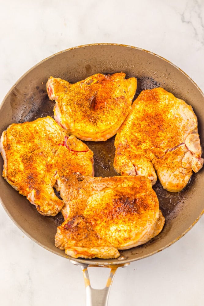 A frying pan with four seasoned raw pork chops on a white marble surface.