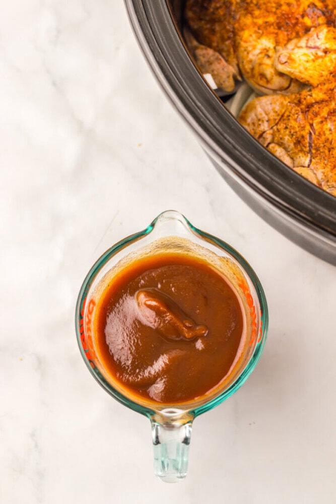 A glass measuring cup filled with barbecue sauce sits on a white surface next to a slow cooker containing seasoned chicken.