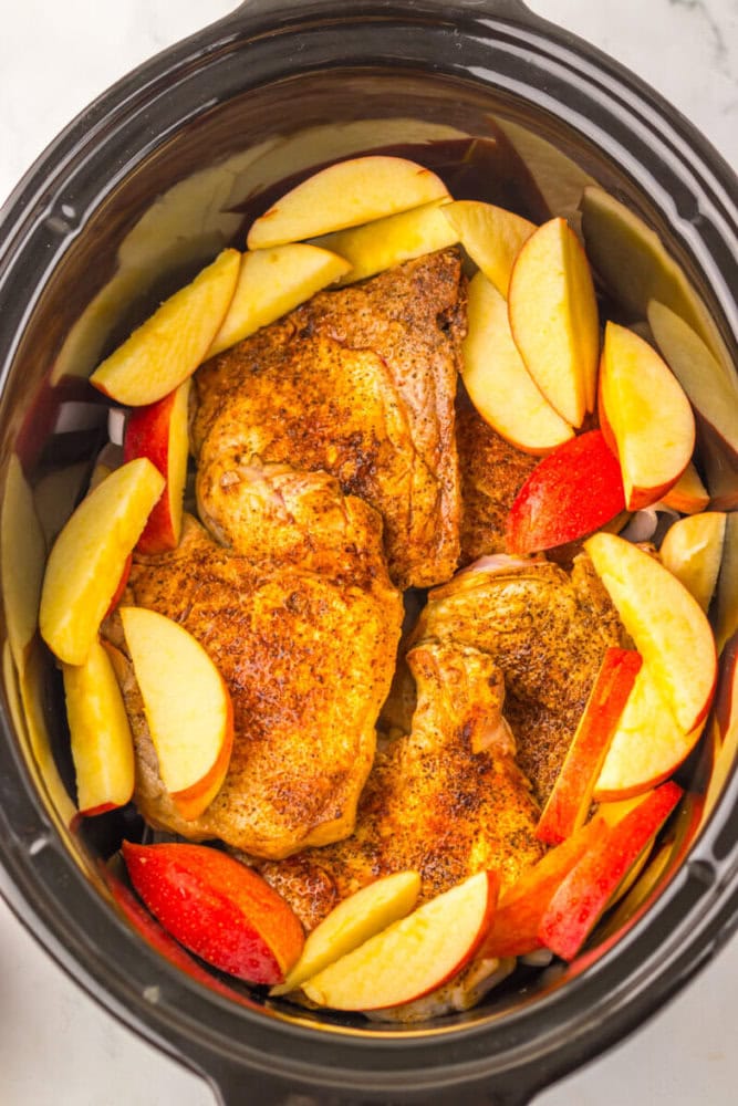 Seasoned chicken thighs and sliced apples arranged in a slow cooker, ready to be cooked.