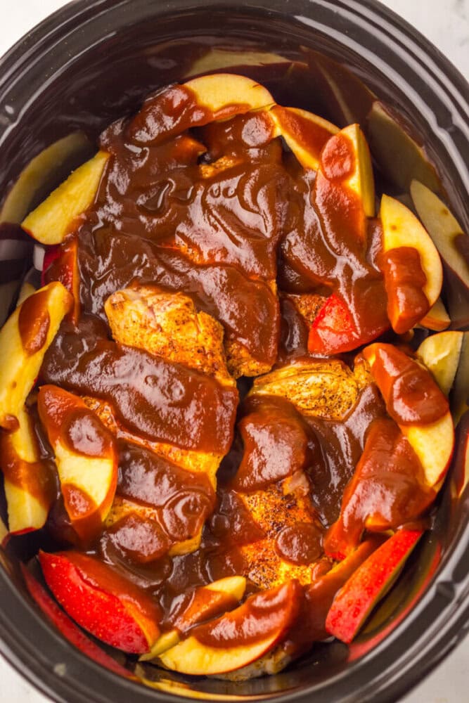 Raw chicken breasts topped with barbecue sauce and surrounded by sliced apples in a slow cooker, ready for cooking.