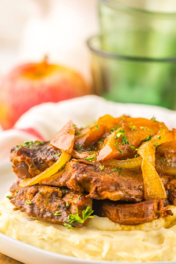 Braised pork chops topped with cooked onions and apples, served over a bed of mashed potatoes on a white plate.