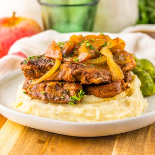 Slices of cooked meat with onions served on mashed potatoes, garnished with herbs, with Brussels sprouts on the side, on a white plate.
