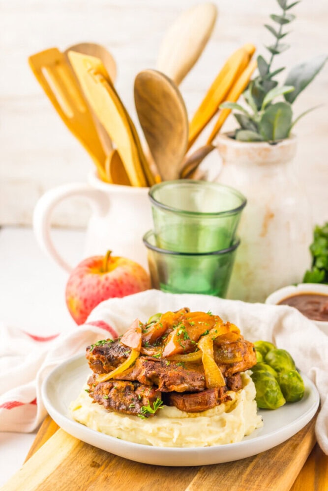 A plate of mashed potatoes topped with cooked meat and onions, served with green vegetables, sits on a wooden board with kitchen utensils and an apple in the background.