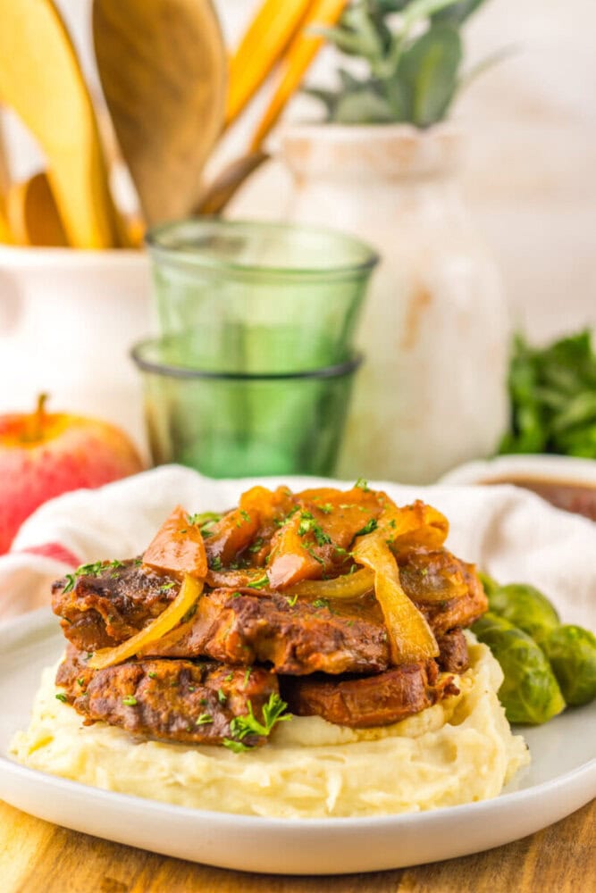 A plate of mashed potatoes topped with braised meat and caramelized onions, served with Brussels sprouts and garnished with herbs.