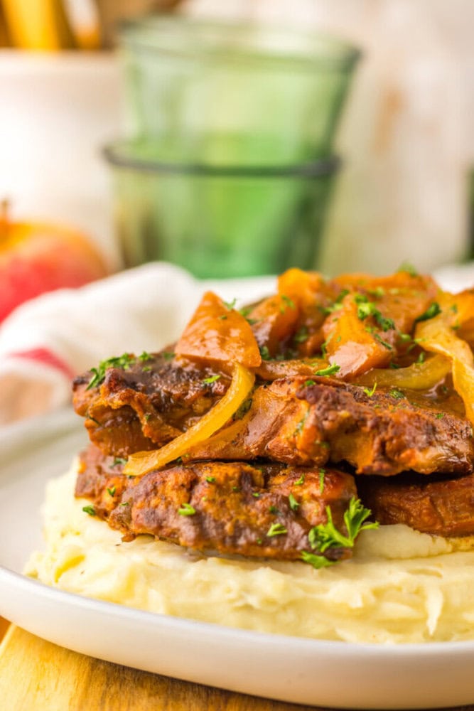 Two pieces of smothered steak with onions and sauce served on a bed of mashed potatoes, garnished with chopped parsley on a white plate.
