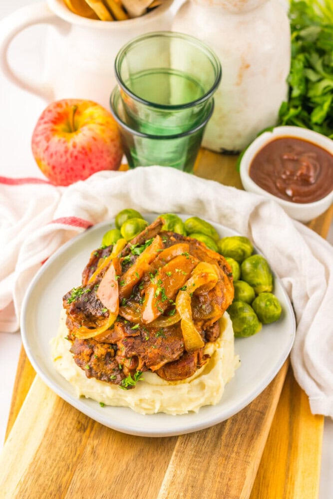 A plate of mashed potatoes topped with braised meat, onions, and gravy, served with Brussels sprouts, on a wooden board with an apple and glasses in the background.