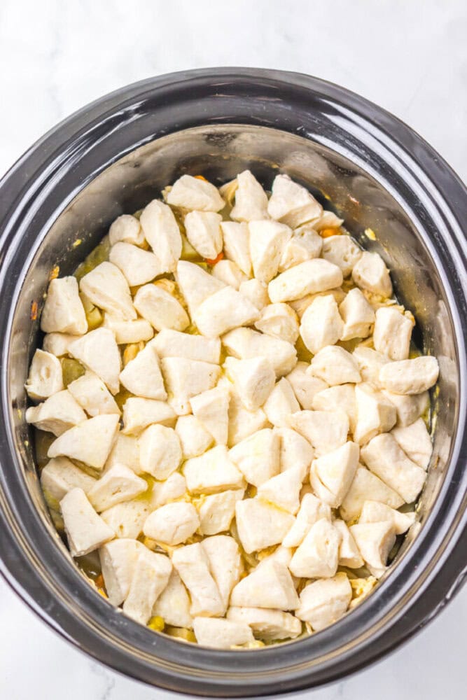 Uncooked biscuit dough pieces spread over a mixture in a black slow cooker, ready to be cooked.