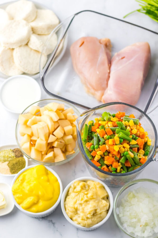 Ingredients for a chicken pot pie recipe arranged on a countertop, including raw chicken breasts, assorted vegetables, biscuit dough, diced potatoes, soups, and seasonings.