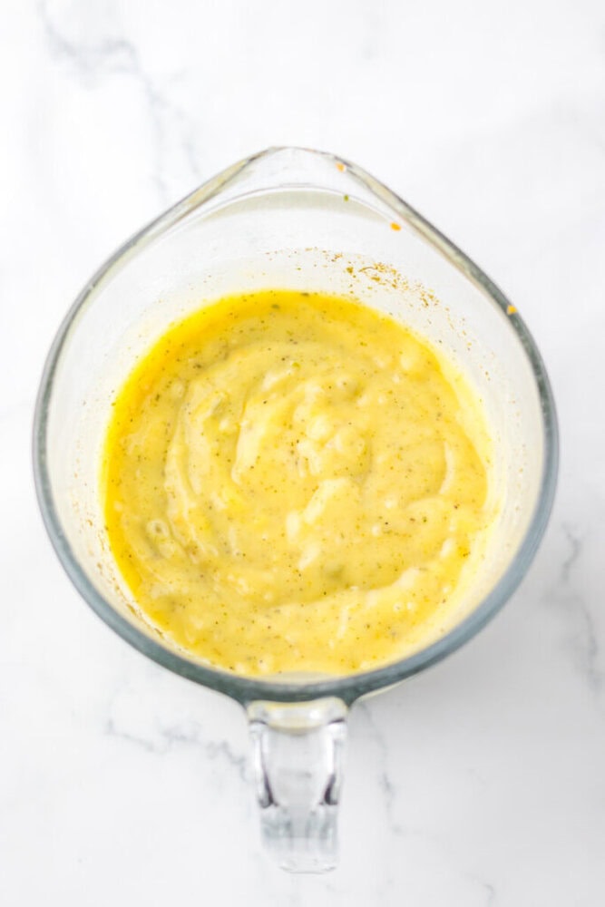 A glass measuring cup filled with a thick, yellow sauce or dressing, placed on a white marble surface.