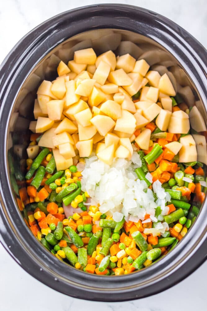 Chopped potatoes, onions, and mixed vegetables inside a slow cooker, ready to be cooked.