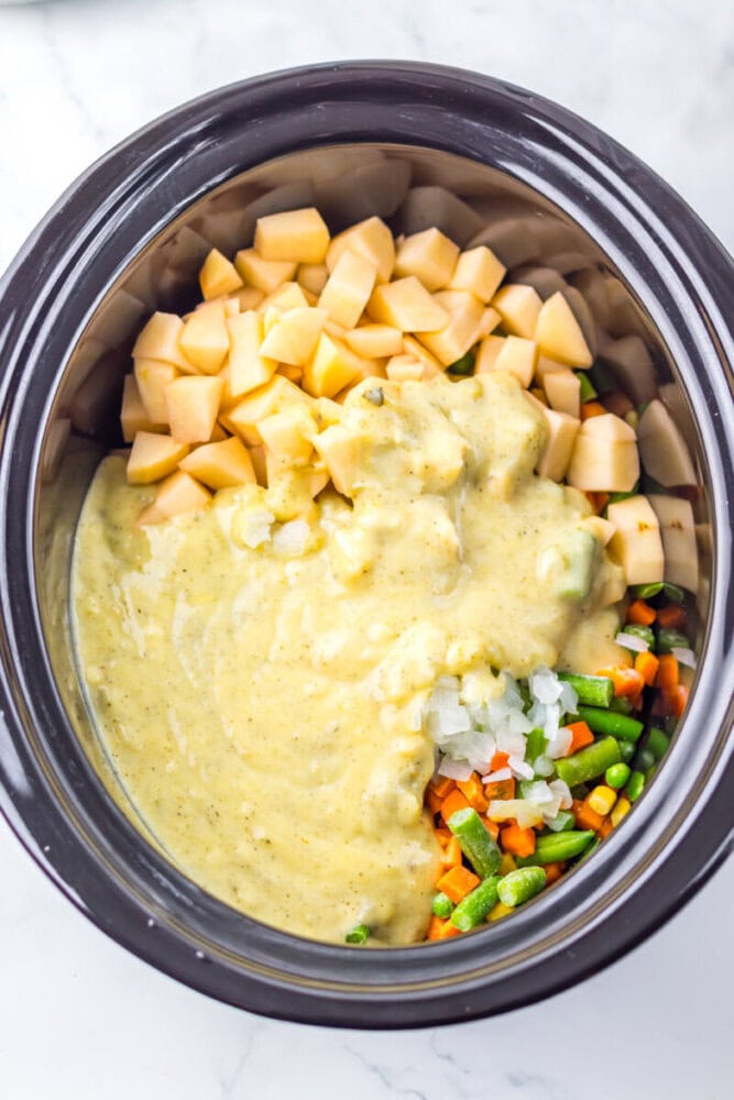 Chopped sweet potatoes, mixed vegetables, diced onions, and a creamy soup mixture in a slow cooker, ready to be cooked.