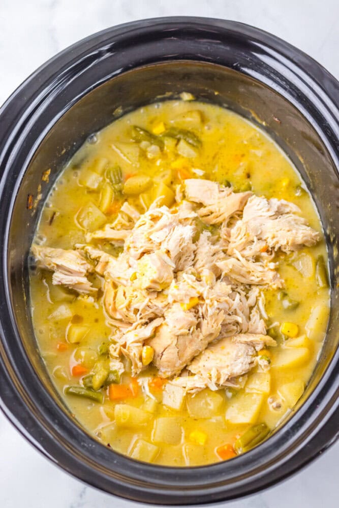 Slow cooker filled with shredded chicken, diced potatoes, mixed vegetables, and yellow broth, viewed from above on a marble surface.