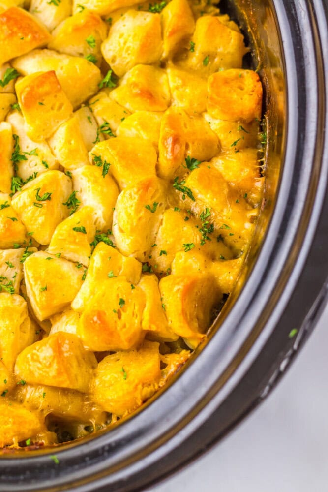 Close-up of golden-brown biscuit pieces baked with melted cheese and herbs in a slow cooker.