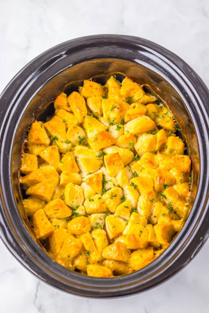 A slow cooker filled with golden, cubed biscuit pieces coated in a creamy, seasoned sauce, garnished with chopped herbs.