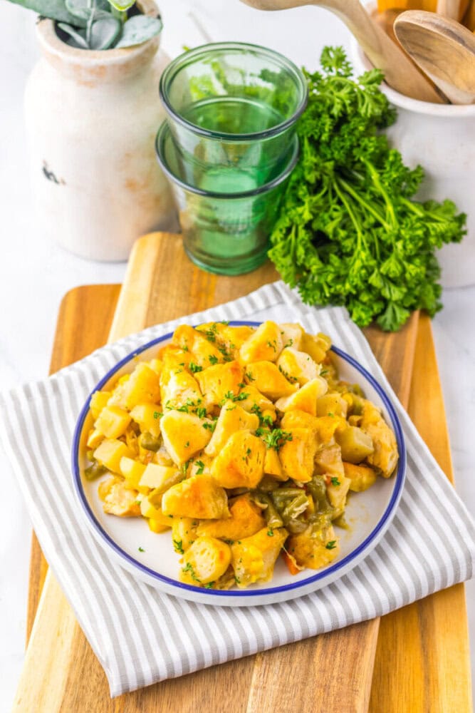 A plate of cooked vegetables and potatoes garnished with herbs sits on a striped cloth, with fresh parsley and glassware in the background.