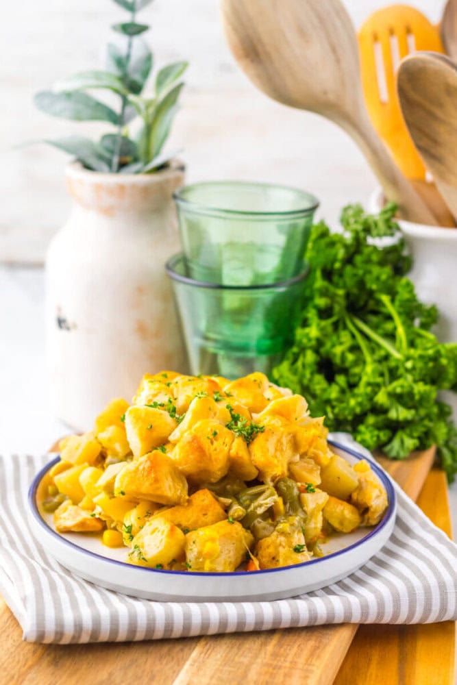 A plate of cooked potato salad with herbs sits on a striped cloth, surrounded by green glasses, wooden utensils, fresh parsley, and a decorative vase in the background.