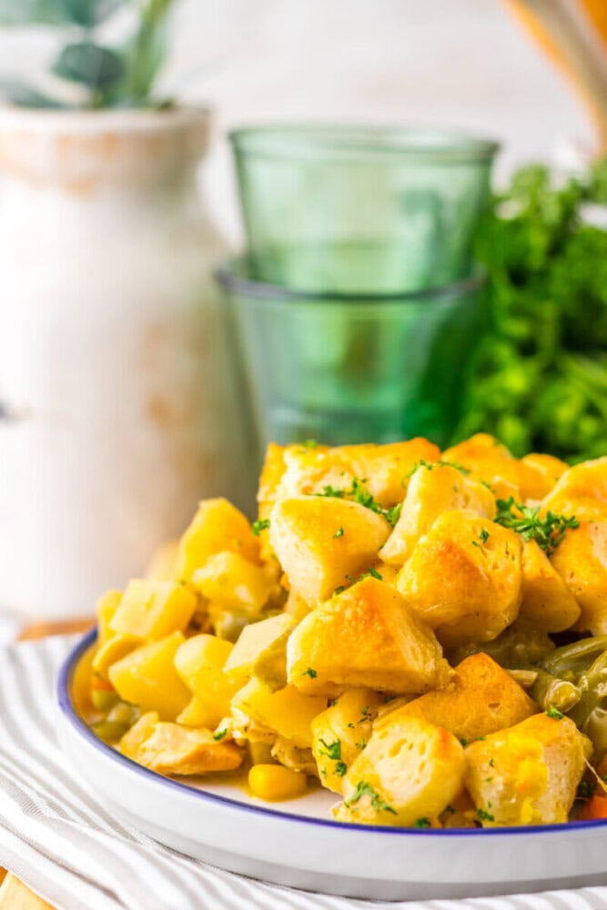 A plate of baked biscuits, potatoes, green beans, and corn garnished with chopped parsley, with glass cups and a white vase in the background.