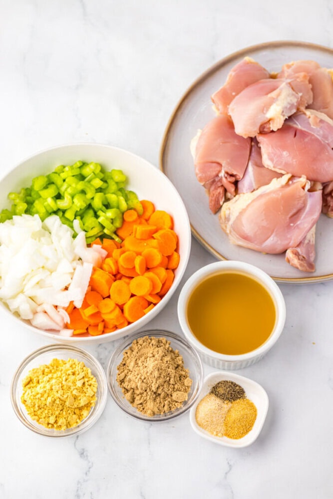 Raw chicken thighs on a plate, and bowls containing chopped onions, celery, carrots, chicken broth, brown sugar, mustard powder, and assorted spices on a white surface.