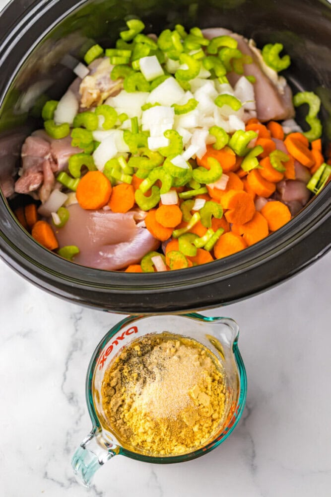 Slow cooker filled with raw chicken, chopped carrots, celery, and onions, next to a measuring cup containing dry seasoning mix.