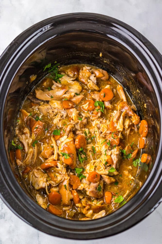A slow cooker filled with shredded meat, sliced carrots, and herbs in a thick sauce, viewed from above.