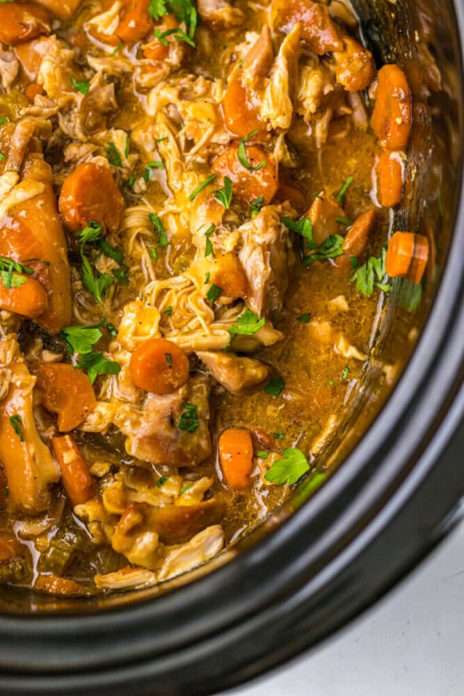 Close-up of shredded chicken, carrots, and herbs in a thick sauce inside a slow cooker.