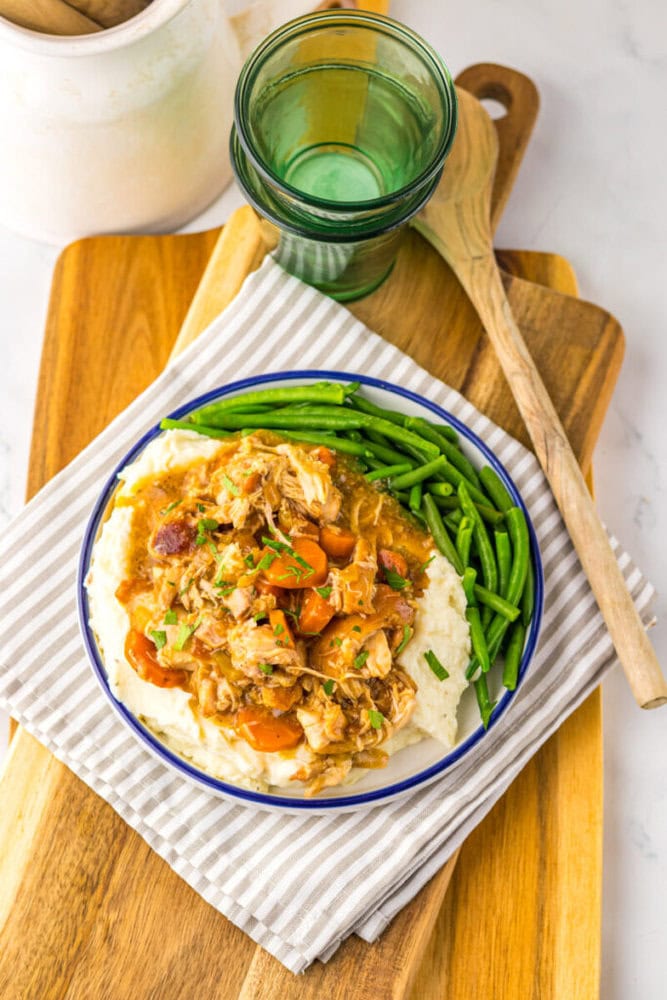 A plate of shredded meat with carrots and gravy served over mashed potatoes, with green beans on the side, sits on a striped napkin next to a green glass and wooden spoon.