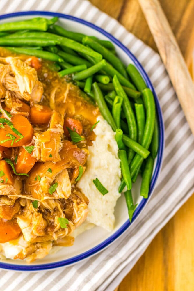 A plate of shredded chicken with carrots and sauce served over mashed potatoes, with a side of green beans.