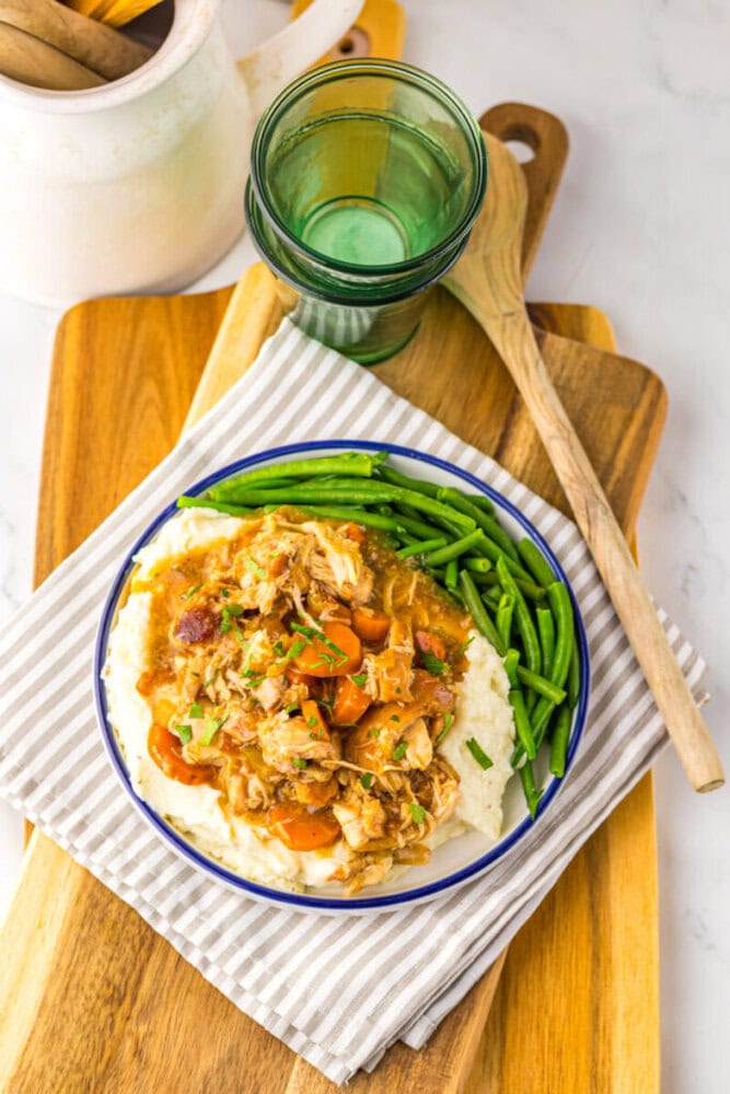 A plate of shredded chicken stew with carrots served over mashed potatoes, alongside green beans, on a striped cloth with a glass, wooden spoon, and layered cutting boards.