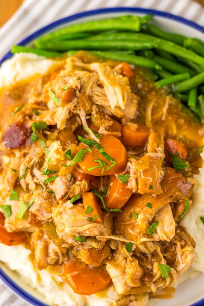 A plate of mashed potatoes topped with shredded chicken, carrots, and gravy, garnished with herbs, with green beans on the side.