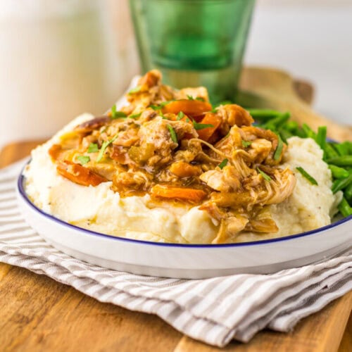 A plate of mashed potatoes topped with shredded chicken and carrots in sauce, served with green beans on the side, sits on a striped napkin and wooden board.