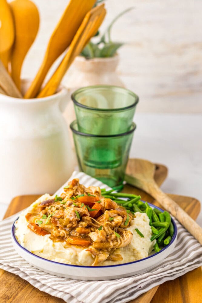 A plate of chicken and vegetables in sauce served over grits with green beans on the side, placed on a wooden board near glasses, utensils, and a striped napkin.
