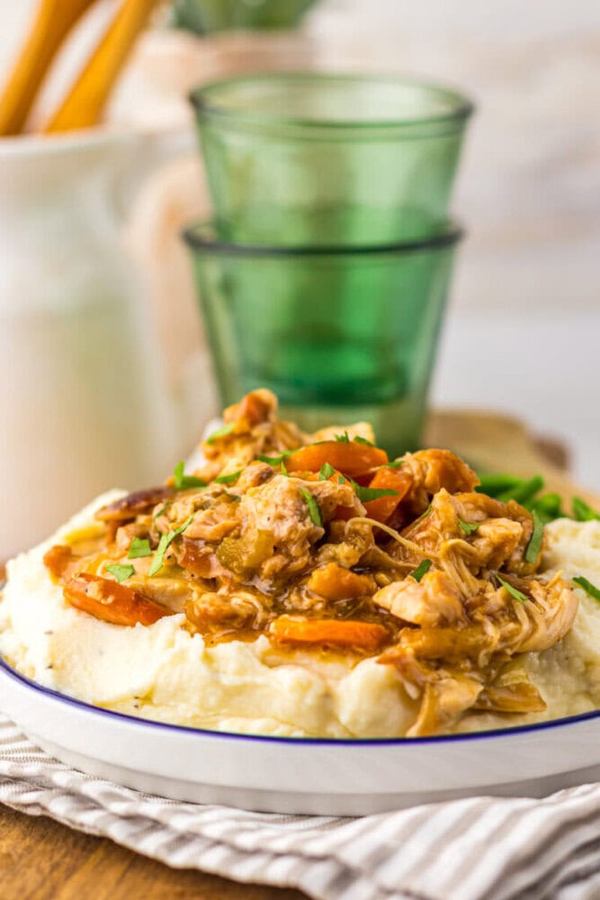 A plate of mashed potatoes topped with shredded chicken, carrots, and gravy, garnished with herbs; green glasses are in the background.