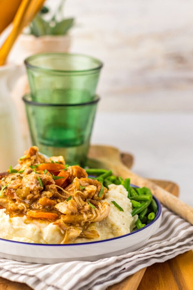 A plate of shredded chicken with gravy served over mashed potatoes, alongside green beans, on a striped napkin with green glass cups in the background.