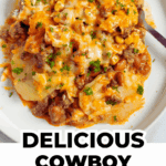 A plate of cowboy casserole topped with melted cheese, beans, corn, ground beef, and chopped parsley, with a fork on the side.