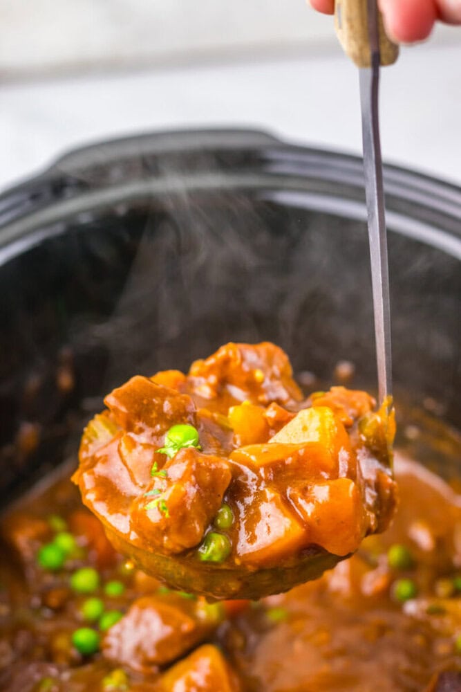 A ladle holding a serving of beef stew with potatoes and peas over a pot, with steam rising from the hot dish.