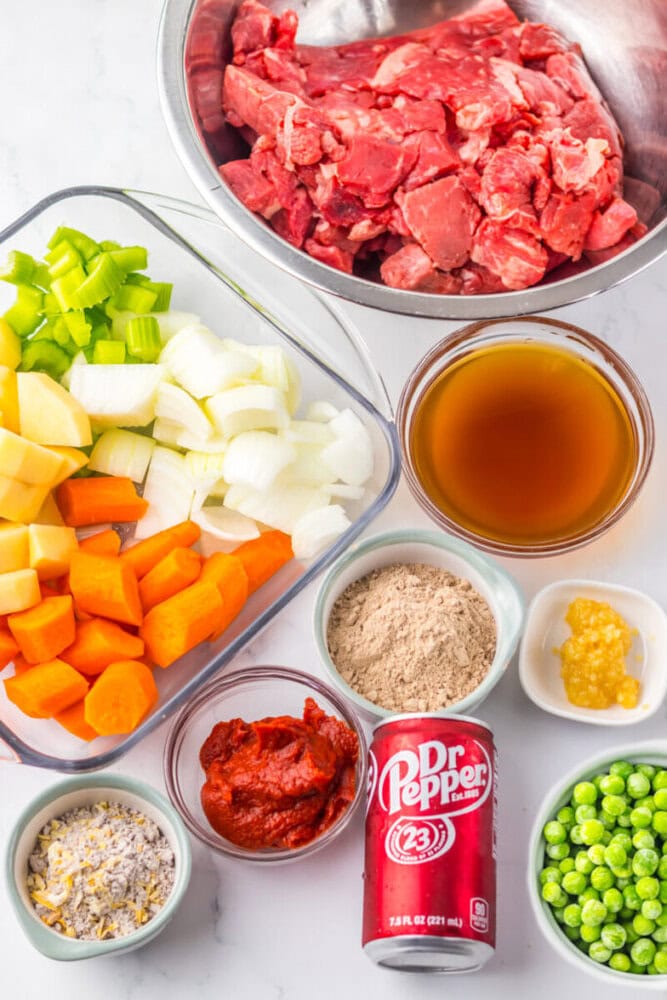 Assorted ingredients for a beef stew recipe, including raw beef, chopped vegetables, beef broth, peas, tomato paste, seasonings, garlic, and a can of Dr. Pepper, arranged on a countertop.