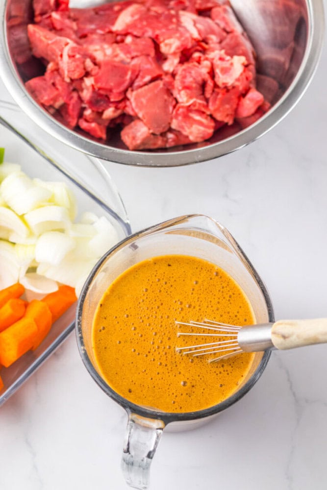 A glass measuring cup with a whisk inside contains a brown-orange sauce. Nearby are bowls of raw beef, chopped onions, and chopped carrots.