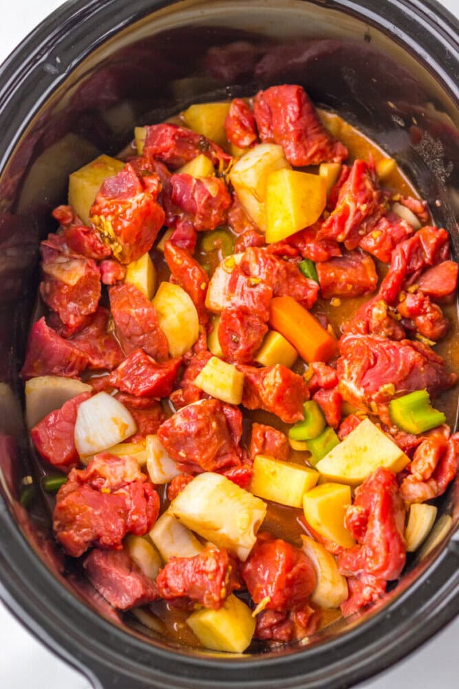 Chunks of raw beef, potatoes, carrots, onions, and celery in a slow cooker with broth, ready to be cooked.